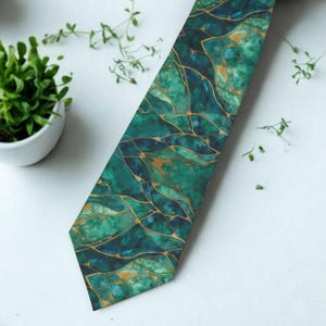 May include: A green and gold patterned necktie. The tie features a watercolor-like design with shades of teal, emerald, and gold. The tie is angled across a white surface, with a small potted plant and sprigs of greenery in the background.