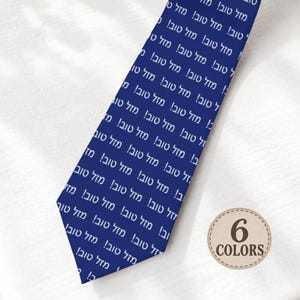 May include: A dark blue necktie featuring a repeating white pattern of the Hebrew text "Mazel Tov". A circular beige badge with a stitched border is positioned in the lower right corner, displaying the text "6 COLORS" in dark brown lettering.