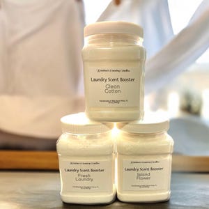 May include: Three clear plastic jars of laundry scent booster. The jars are stacked on top of each other. The top jar is labeled "Clean Cotton", the middle jar is labeled "Fresh Laundry", and the bottom jar is labeled "Island Flower".
