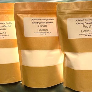 May include: Three brown paper bags with white labels. Each bag contains a white powder and is labeled with the text "Kristina's Country Candles Laundry Scent Booster" followed by a scent name: "Ocean Waves", "Clean Cotton", and "Fresh Laundry".