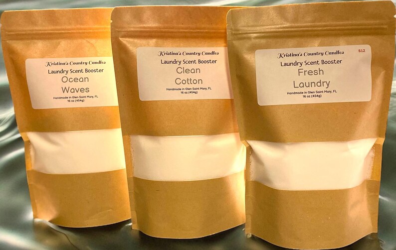 May include: Three brown paper bags with white labels. Each bag contains a white powder and is labeled with the text "Kristina's Country Candles Laundry Scent Booster" followed by a scent name: "Ocean Waves", "Clean Cotton", and "Fresh Laundry".