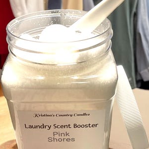 May include: A clear plastic jar filled with white laundry scent booster crystals. The jar has a white label with the text "Kristina's Country Candles Laundry Scent Booster Pink Shores Handmade in Glen Saint Mary, FL 32 oz (907g)".