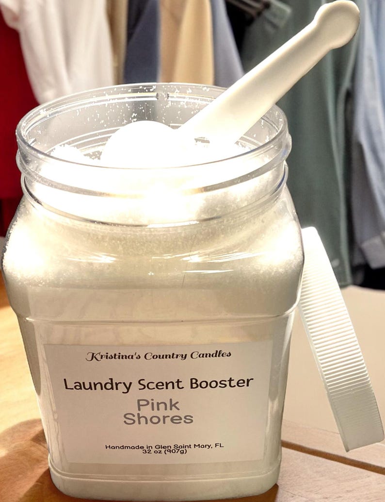 May include: A clear plastic jar filled with white laundry scent booster crystals. The jar has a white label with the text "Kristina's Country Candles Laundry Scent Booster Pink Shores Handmade in Glen Saint Mary, FL 32 oz (907g)".