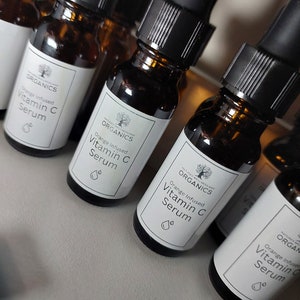 K&ouml;nnte beinhalten: Mehrere bernsteinfarbene Glasflaschen mit schwarzen Tropfern und wei&szlig;en Etiketten. Die Etiketten zeigen ein Baumlogo und den Text "NATURE'S WELLNESS AND ORGANICS" und "Orange Infused Vitamin C Serum".