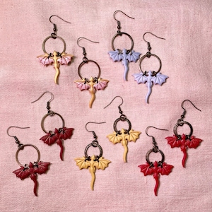 May include: Ten pairs of dangle earrings featuring a dragon design. The earrings are made of metal and have a variety of colors, including red, yellow, and purple.