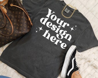 May include: A dark gray t-shirt with the text "Your design here" in white. The shirt is laying on a white fuzzy surface with a brown leather purse and black and white sneakers.