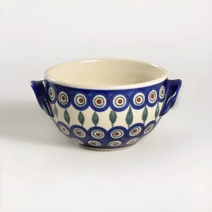 May include: A ceramic bowl with two handles, featuring a cream-colored interior and a blue exterior adorned with a repeating pattern of red, green, and white circles and teardrop shapes. The bowl is designed for serving soups or stews.