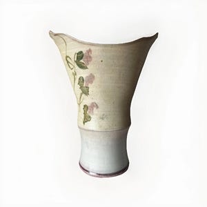 May include: A handcrafted ceramic vase with a wide, flaring top and a cylindrical base. The vase features a cream-colored body with delicate pink floral and green leaf accents. The base is a light blue-gray color.
