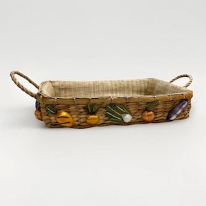 May include: Rectangular woven basket with two handles. The basket is lined with a tan fabric and decorated with colorful, three-dimensional fruit and vegetable embellishments, including lemons, onions, and eggplants. The basket is designed for decorative or storage use.