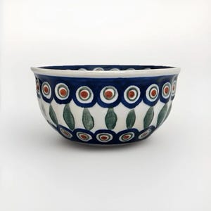 May include: A ceramic bowl with a white interior and a blue rim. The exterior features a repeating pattern of blue circles with red centers, green leaf shapes, and white background. The bowl is likely used for serving food.