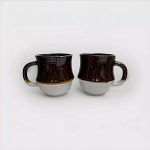 May include: Two handmade ceramic mugs. The top half of each mug is a dark brown color, with a gold band separating it from the light gray bottom. Each mug has a curved handle.