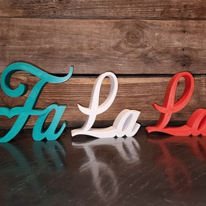 Puede incluir: Decoración navideña de madera « Fa La La » en color turquesa, blanco y rojo. Las letras cursivas se colocan sobre un fondo de madera rústica. El « Fa » turquesa tiene un corazón. Ideal para la decoración festiva.
