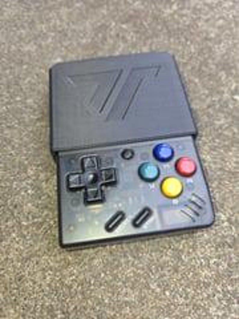 Puede incluir: Una consola de juegos port&aacute;til negra con una pantalla abatible. La consola cuenta con un mando direccional, botones de acci&oacute;n de colores y un logotipo en la cubierta superior. Dise&ntilde;ada para juegos port&aacute;tiles.