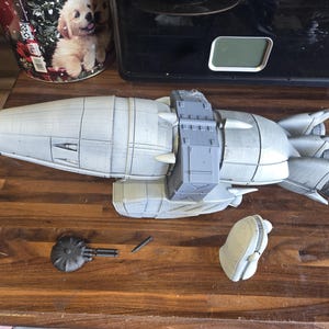 May include: A 3D printed grey model of a spaceship with a tapered nose, a central rectangular section, and three rear thrusters. Included are two separate smaller components: a circular piece with two thin rods and a curved, helmet-like object.