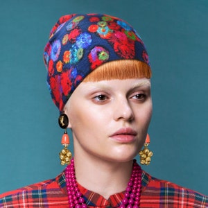 以下が含まれることがあります： 赤と青の鮮やかな模様のヘッドスカーフ、ピンクのビーズネックレス、コーラルとゴールドのイヤリングをつけた女性。彼女は赤と黒のチェックシャツを着ています。