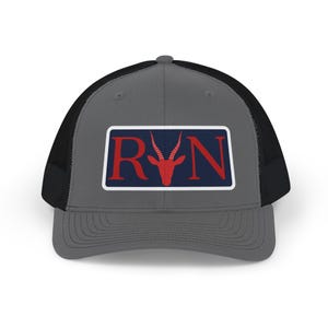 Puede incluir: Gorra de camionero gris con parte trasera de malla negra. La parte delantera presenta un parche rectangular con las letras "RVN" en rojo, con una silueta de cabeza de antílope roja, sobre un fondo azul marino, enmarcado por un borde blanco.