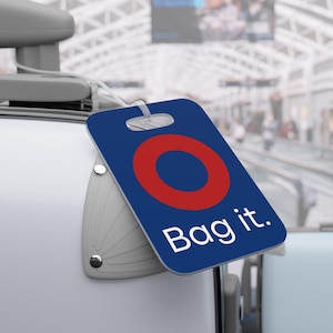 May include: A blue luggage tag with a red circle design and the words "Bag it." in white. The tag is attached to a white suitcase, suggesting travel and luggage.