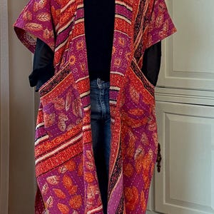 May include: A long, open-front kimono-style jacket in a patchwork design. The jacket features a vibrant mix of colors, including purple, orange, and white, with leaf and geometric patterns. It has wide sleeves and two front pockets. The garment is worn over a black top and blue jeans.