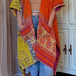 May include: A long, open-front kimono jacket crafted from quilted fabric featuring a vibrant patchwork of orange, yellow, and pink floral patterns. The garment includes two large front patch pockets and visible white running stitch detailing.