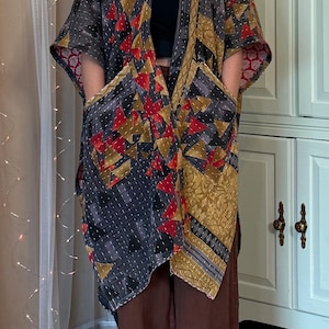 May include: A kimono-style jacket featuring a patchwork design of geometric triangles in shades of red, gold, and black. The garment has short, wide sleeves, an open front, and two large patch pockets on the front, accented with visible white stitching.