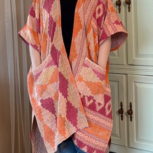 Könnte beinhalten: Eine farbenfrohe, offen geschnittene Kimono-Jacke mit einem geometrischen Muster in Orange-, Rosa- und Pfirsichtönen. Die Jacke hat große Taschen und eine dekorative Bordüre. Das Kleidungsstück wird über einem schwarzen Oberteil und einer blauen Jeans getragen.