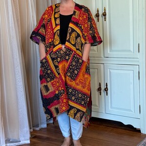 May include: A knee-length open-front kimono jacket featuring a patchwork design with intricate floral and paisley patterns in shades of red, orange, yellow, and black. The garment includes short sleeves and two large patch pockets on the front.
