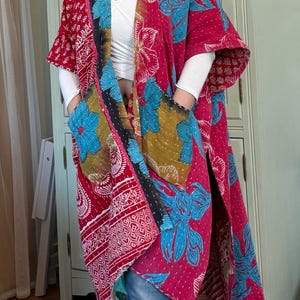 Op de afbeelding: Een lange, open kimono-achtige robe in tinten rood, roze, blauw en geel. De robe heeft grote bloempatronen en patchwork-ontwerpen. Het heeft zakken en is gemaakt van een lichte stof, geschikt om in laagjes te dragen.