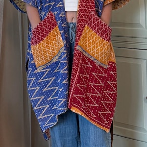 Könnte beinhalten: Eine gesteppte Jacke im Kimono-Stil mit einem Patchwork-Design aus blauem, rotem und gelbem Stoff mit weißen Zickzack-Nähten. Das Kleidungsstück verfügt über zwei große Vordertaschen und kurze Ärmel und wird über einem weißen Oberteil und blauen Jeans getragen.