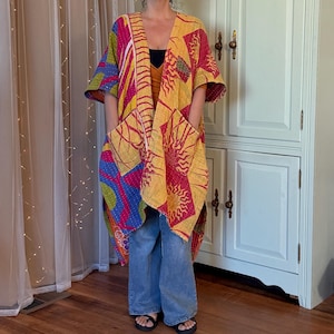 May include: A long, open-front kimono-style jacket featuring a vibrant patchwork design with yellow, pink, and blue sun motifs and geometric patterns. The garment has wide, short sleeves, a relaxed fit, and two large patch pockets on the front.