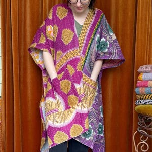 May include: A vibrant, knee-length kimono-style robe in shades of purple, yellow, and green. The robe features a patchwork design with floral and geometric patterns, and two front pockets. The garment is worn by a person.