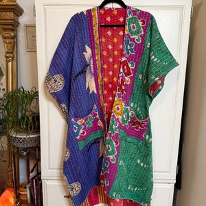May include: A vibrant, quilted kimono jacket with a patchwork design. The jacket features blue, green, pink, and red fabrics with floral and geometric patterns. It has wide sleeves and two front pockets, hanging on a black hanger.