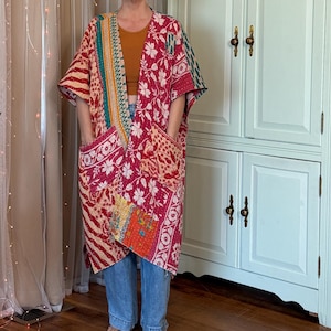 May include: A long, open-front kimono-style jacket featuring a patchwork design of various floral and geometric patterns in shades of red, pink, orange, and teal. The garment includes two large front pockets and is constructed with visible stitching.