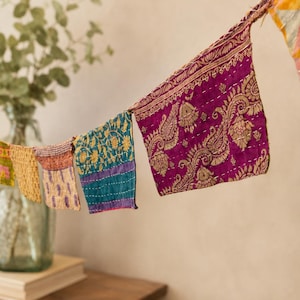 Puede incluir: Una guirnalda decorativa que presenta una serie de pequeñas banderas rectangulares de tela hechas de textiles reutilizados. Cada bandera muestra patrones y colores distintos, incluyendo magenta intenso, verde azulado y amarillo, con costuras blancas visibles.