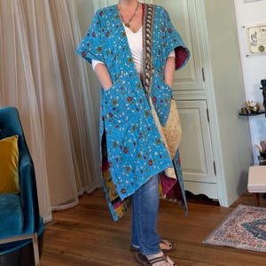 May include: A long, open-front quilted duster coat in a vibrant turquoise blue fabric featuring a small-scale floral pattern in red, yellow, and green. The garment includes two large patch pockets on the front and a contrasting dark patterned trim.