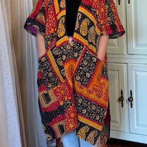 Pode incluir: Um casaco tipo quimono aberto à frente, pelo joelho, com um design de patchwork de padrões florais e paisley em tons de vermelho, laranja, amarelo e preto. A peça tem mangas curtas, dois bolsos frontais grandes e costuras brancas visíveis.