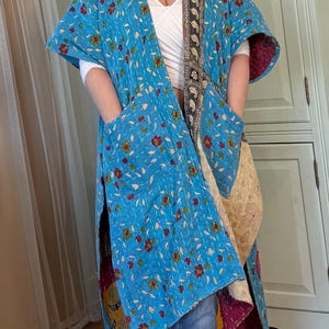 Op de afbeelding: Lichtblauwe kimono-jas met een bloemenpatroon in rood, geel en groen. De jas heeft een gewatteerde textuur en een contrasterende patroonrand in zwart, goud en crème. Het heeft grote zakken en een bordeauxrode voering.