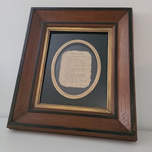 May include: A framed piece of paper with handwritten text, set within a dark wooden frame. The frame has a black inner border and a gold oval frame around the paper. The text is in a cursive script.