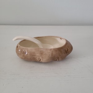 May include: A ceramic bowl shaped like a brown potato with a textured, uneven surface and a smooth, cream-colored interior. A small, matching cream-colored ceramic spoon rests inside the bowl, extending over the side of the potato-shaped rim.
