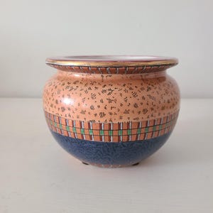 May include: A small, round ceramic pot with a wide opening. The pot features a peach-colored upper section with a dotted pattern, a plaid band in orange, green, and white, and a navy blue base. The rim is trimmed with gold.