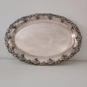 May include: Oval silver-toned serving tray with a decorative border of grape clusters and leaves. The tray has a smooth, reflective surface and is likely made of metal. A classic piece for serving food or displaying items.
