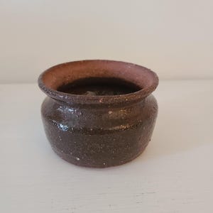 May include: A small, handcrafted, dark brown ceramic pot with a wide, slightly flared rim. The pot has a rounded body and a textured surface, suggesting a rustic or artisanal aesthetic. The interior is a darker shade of brown.
