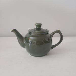 May include: A dark green ceramic teapot with a matching lid and handle. The teapot has a rounded body, a curved spout, and a small knob on the lid. The teapot is sitting on a white surface.