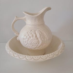 May include: Cream-colored ceramic pitcher and basin set. The pitcher has a curved handle and spout, with a decorative embossed design. The basin has a scalloped edge. The set is speckled with a matte finish.