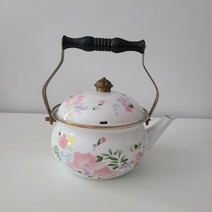 May include: White enamel teapot with a floral design in pink, yellow, and purple. The teapot has a black handle and a brass-colored lid and accents. The spout is white, and the handle is arched.