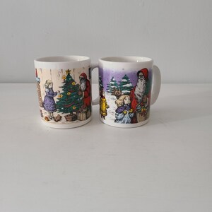 May include: Two white ceramic mugs with Christmas-themed illustrations. One mug depicts a girl decorating a Christmas tree with Santa Claus. The other mug shows children and Santa Claus in a snowy scene. Both mugs have a handle.