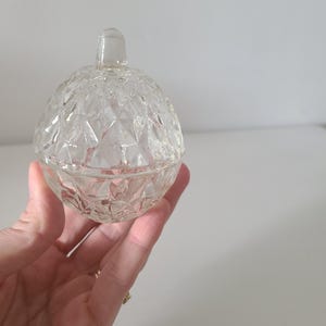 May include: A clear, round glass trinket dish with a lid. The dish and lid are decorated with a diamond pattern. The lid has a small, rounded handle. The dish is held in a hand against a plain white background.