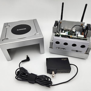 May include: A silver GameCube console with the top removed, revealing the internal components. The console has two antennas and a black power adapter. The console is labeled "DOL-001 PC".