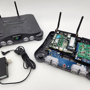 May include: A black Nintendo 64 console with the top removed, revealing the internal components. The console has two antennas and is connected to a black power adapter. The internal components include a green circuit board with wires and a blue circuit board with a red button.