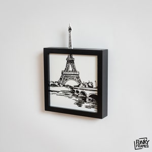 Pode incluir: Arte de parede com moldura preta apresentando uma ilustração em preto e branco da Torre Eiffel e uma ponte sobre um rio. Uma Torre Eiffel em miniatura se estende da parte superior da moldura. A obra de arte é montada em uma parede branca.