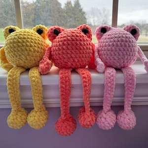 May include: Three crocheted frog figurines with long legs. The frogs are sitting on a windowsill. The frog on the left is yellow, the middle frog is orange, and the frog on the right is pink.
